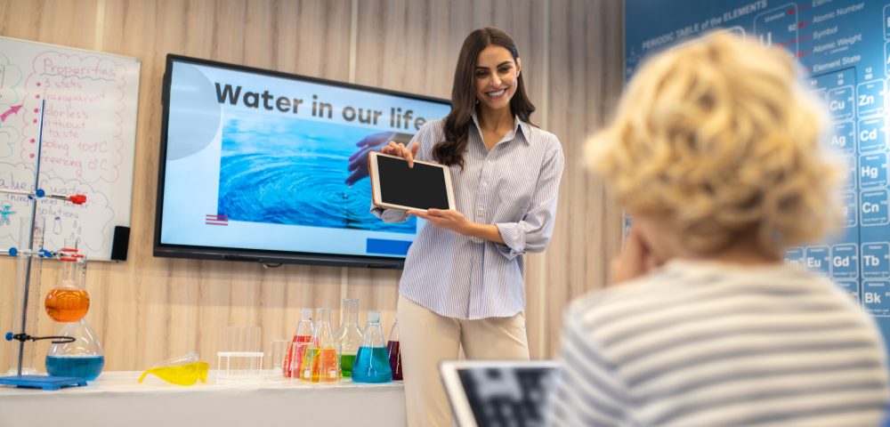 Important information. Young smiling standing woman pointing finger at tablet screen showing students sitting back to camera at desks